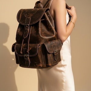 May include: A brown leather backpack with a distressed finish. The backpack features a flap closure, drawstring, and two front pockets with buckle closures. The bag has adjustable shoulder straps and a top handle. The leather has a textured appearance.