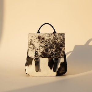 Puede incluir: Una mochila cuadrada con un estampado de piel de vaca en blanco y negro. La bolsa tiene un asa negra, correas negras y hebillas de color latón. La mochila está sobre una superficie blanca.