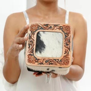 May include: Square leather box with a floral tooled design and a cowhide center. The box features a light brown border with a dark gray background and a black and white cowhide center. The box is held by a person.