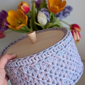 Pode incluir: Uma cesta feita &agrave; m&atilde;o em tons de lavanda e azul claro, com uma tampa de madeira e uma pequena al&ccedil;a arredondada. A textura da cesta &eacute; vis&iacute;vel. Um buqu&ecirc; de tulipas coloridas e outras flores est&aacute; ao fundo.