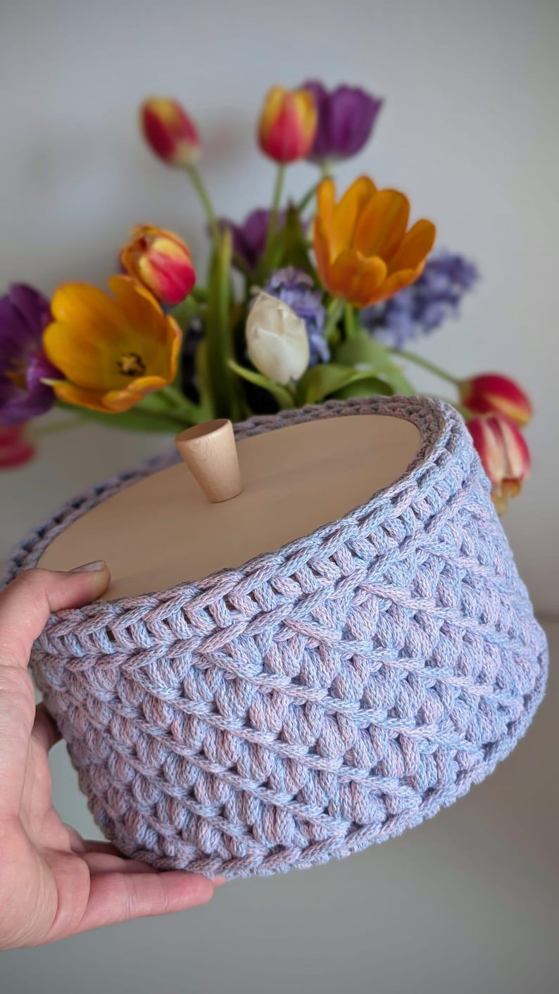 Pode incluir: Uma cesta feita &agrave; m&atilde;o em tons de lavanda e azul claro, com uma tampa de madeira e uma pequena al&ccedil;a arredondada. A textura da cesta &eacute; vis&iacute;vel. Um buqu&ecirc; de tulipas coloridas e outras flores est&aacute; ao fundo.
