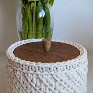 Pode incluir: Uma cesta redonda de croch&ecirc; creme com tampa de madeira e um pequeno bot&atilde;o de madeira. A cesta tem um padr&atilde;o tecido. Um vaso de flores est&aacute; ao fundo.
