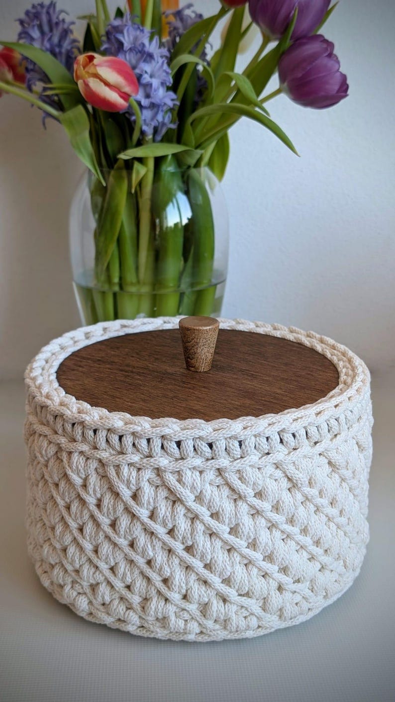 Pode incluir: Uma cesta redonda de croch&ecirc; creme com tampa de madeira e um pequeno bot&atilde;o de madeira. A cesta tem um padr&atilde;o tecido. Um vaso de flores est&aacute; ao fundo.