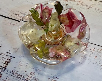 May include: A clear resin ring holder with dried flowers and leaves embedded inside. The holder is shaped like a flower with a raised center for the ring.