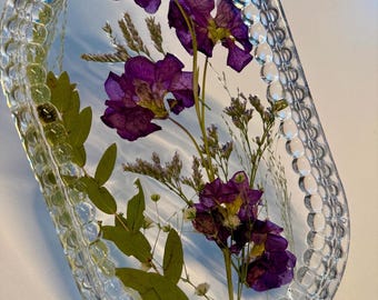 May include: Oval-shaped clear resin tray with a decorative border. The tray is embedded with dried purple flowers, green leaves, and small white flowers. The resin is transparent, allowing the floral arrangement to be fully visible.