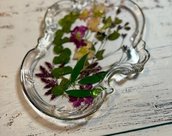 May include: A clear glass tray with a decorative, ornate edge. The tray is filled with pressed flowers and leaves, including pink, white, and green hues.