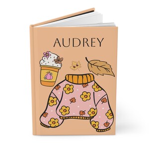 Puede incluir: Un diario con el nombre "AUDREY" en la portada. La portada presenta ilustraciones de una bebida de café, un suéter con flores amarillas y hojas de otoño en tonos de amarillo, naranja y rosa.