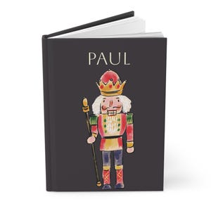 Puede incluir: Un diario de tapa dura negro con el nombre "PAUL" en blanco. La portada presenta una ilustración de acuarela de un soldado cascanueces con corona y bastón. El cascanueces es rojo, amarillo y azul.