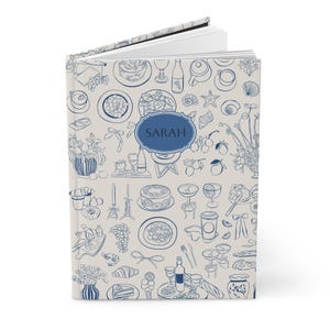 May include: A cream-coloured journal with a blue and white pattern of food and drink illustrations. The journal has the name "SARAH" printed on the front cover.