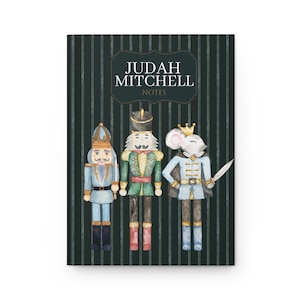 Pode incluir: Um caderno verde-escuro com riscas brancas apresentando o texto JUDAH MITCHELL NOTES numa moldura decorativa. Abaixo do texto, três figuras em estilo aguarela: um quebra-nozes azul, um verde e um rei rato com uma espada.