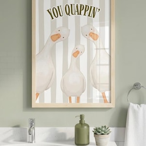 May include: A framed wall art print featuring three white geese with orange beaks against a background of vertical beige and white stripes. The text "YOU QUAPPIN'" is printed in dark green letters at the top center of the illustration.
