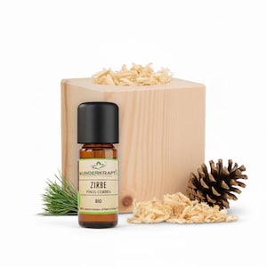 Peut inclure: Une petite bouteille en verre ambré avec un bouchon noir, étiquetée "Zirbe Pinus Cembra BIO", est placée devant un bloc de bois rempli de copeaux. Une pomme de pin et des aiguilles de pin sont également présentes. Il s'agit probablement d'une huile essentielle.