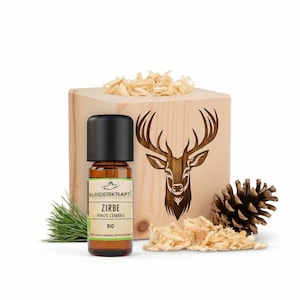 Peut inclure: Une petite bouteille en verre ambré avec un bouchon noir, étiquetée "Zirbe" et "Pinus Cembra BIO", est placée devant une boîte en bois avec un cerf sculpté. La boîte est remplie de copeaux de bois, avec une pomme de pin et une brindille de pin.