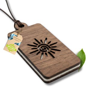 Peut inclure: Pendentif en bois avec un motif de soleil découpé au centre. Le pendentif est rectangulaire avec un cordon en cuir marron. Une étiquette indique "Handgemacht in Bayern" et "NATURAL". Une feuille verte est en arrière-plan.