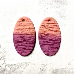 May include: Two oval-shaped earrings with a textured surface. The top half of each earring is a light peach color, transitioning to a deep purple at the bottom. Each earring has a small hole at the top for hanging.