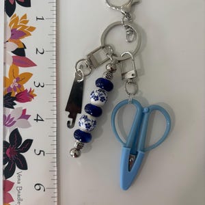 May include: A silver-toned metal keychain featuring a light blue plastic thread cutter, a silver metal charm, and a strand of beads. The beads alternate between solid dark blue and white with blue floral patterns. A ruler shows the item is 5 inches long.