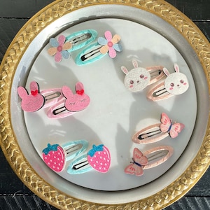 May include: A collection of colorful hair clips featuring various designs, including butterflies, strawberries, bunnies, and flowers. The clips are displayed on a gold-rimmed plate.