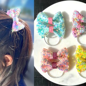 May include: Four pairs of colorful sequined hair bows with silver elastic bands. The bows are in shades of pink, blue, yellow, and orange.