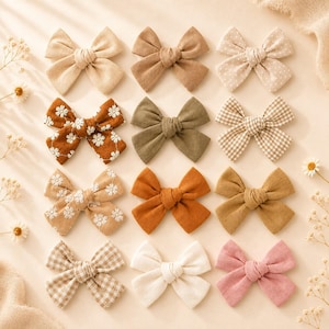 May include: A collection of fabric hair bows in various colors and patterns. The bows include solid colors like beige, brown, and pink, as well as patterned bows with polka dots, gingham, and floral designs. The bows are arranged on a light-colored surface.