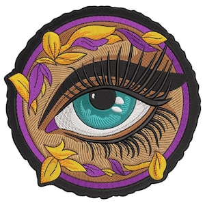 Puede incluir: Parche bordado con un ojo detallado, largas pestañas negras y un iris turquesa. El ojo está rodeado por un diseño floral morado y dorado, sobre un fondo marrón y un borde negro. Ideal para ropa o accesorios.