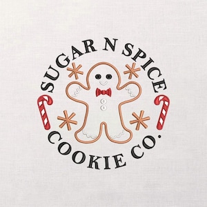May include: Embroidered design featuring a gingerbread man with a red bow tie, surrounded by snowflakes and candy canes. The text "SUGAR N SPICE COOKIE CO." is in a circular pattern around the gingerbread man.