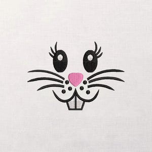 Könnte beinhalten: Gesticktes Hasengesicht mit schwarzen Augen, Wimpern, Schnurrhaaren und einer rosa Nase. Das Hasengesicht ist auf einem weißen Stoffhintergrund gestickt. Das Design zeigt einen lächelnden Ausdruck und eignet sich für Oster- oder Frühlingsmotive.