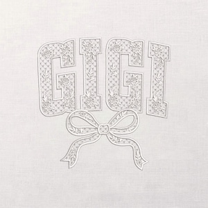 May include: A white textile with the name "GIGI" in a floral-patterned font, with a decorative bow beneath the letters. The letters are filled with a delicate floral design, and the bow has detailed ribbon elements.