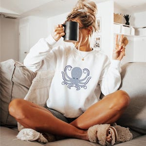 May include: White sweatshirt with a light blue octopus design. The long-sleeved top is worn by a person sitting on a grey sofa, holding a black mug. The person is also wearing grey shorts and fluffy slippers.