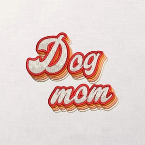 May include: Embroidered patch with the words "Dog mom" in a retro font. The text is white with a red outline and layered orange and pink accents, set against a white fabric background.