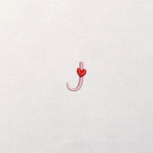 May include: Embroidered letter "j" in a light pink color, with a red heart accent. The design is stitched on a white fabric background. The letter has a curved shape, and the heart is positioned at the top.
