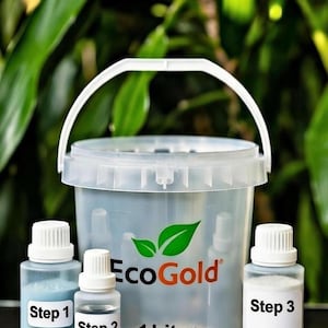May include: A clear plastic bucket with a white handle and the EcoGold logo, accompanied by three small bottles labeled "Step 1", "Step 2", and "Step 3". The bucket is labeled "1 Liter". The background is blurred green foliage.