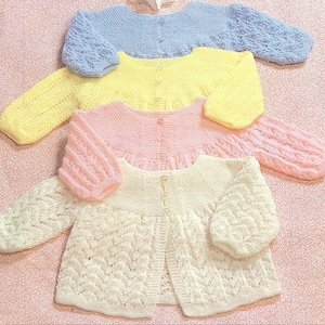 May include: Four knitted baby cardigans in different colours and patterns. The cardigans are laid out on a pink and white floral background. The cardigans are all made with a delicate stitch and have a button fastening.