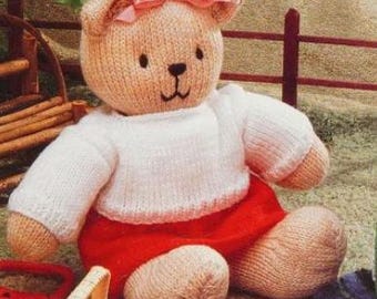 May include: A knitted teddy bear wearing a white sweater and a red skirt. The bear has a pink bow on its head and brown eyes.