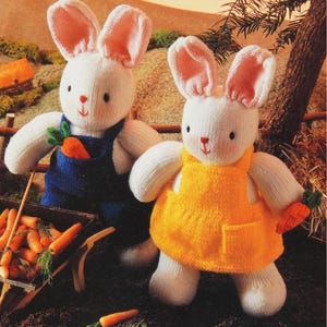 May include: Two white knitted bunny rabbits wearing overalls and a yellow dress, holding carrots. The bunny in overalls is holding a carrot in its hand and the bunny in the yellow dress is holding a carrot in its paw. The bunnies are sitting on a brown surface with green grass and a wooden wheelbarrow filled with carrots.
