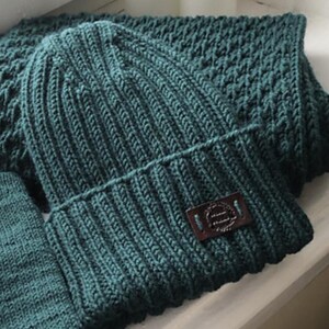 Könnte beinhalten: Eine dunkelgrüne Strickmütze mit einem Lederpatch, auf dem "The Handmade Hat Co." steht.