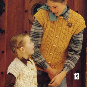 May include: Two girls wearing knitted vests with button closures. The older girl is wearing a yellow vest with wooden buttons and a plaid shirt. The younger girl is wearing a white vest with wooden buttons and a brown shirt. The girls are standing in front of a wooden door.