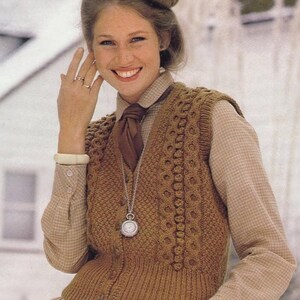May include: A brown knitted vest with a button closure and a cable knit pattern. The vest is worn over a white and brown plaid shirt with a brown tie. The person is wearing a silver necklace with a pocket watch pendant.
