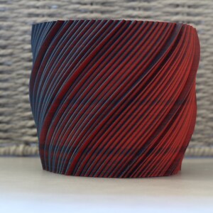 Puede incluir: Un macetero de cerámica rojo y negro con un diseño en espiral. El macetero está hecho de un material texturizado y tiene un fondo liso y redondeado.