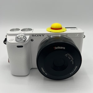 May include: A white Sony camera with a 7Artisans 35mm 1:5.6 lens attached. The camera has a yellow and red hat on top.