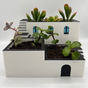 Pode incluir: Vaso de cerâmica branco em forma de casa moderna com vários níveis e detalhes em preto. O vaso está cheio de plantas suculentas.