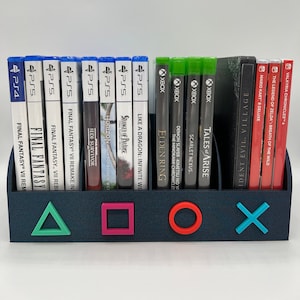 May include: A black video game storage organizer with a blue, green, pink, and turquoise PlayStation and Xbox controller symbols. The organizer holds multiple video game cases, including Final Fantasy VII Remake, Horizon Forbidden West, Elden Ring, Tales of Arise, and Demon Slayer - Kimetsu no Yaiba - The Hinokami Chronicles.