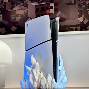 Puede incluir: Una consola PlayStation 5 blanca con una unidad de disco negro se muestra en un soporte blanco en forma de cristal. La consola está iluminada desde abajo con una luz azul.