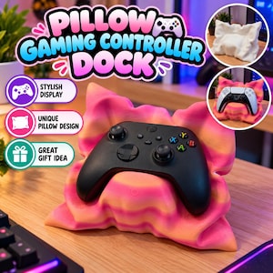 May include: A pink and orange gradient pillow-shaped gaming controller dock holding a black wireless game controller. The dock features a soft, rippled texture resembling a plush cushion. Text on the image reads PILLOW GAMING CONTROLLER DOCK.