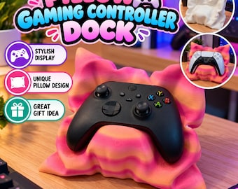 May include: A pink and orange gradient pillow-shaped gaming controller dock holding a black wireless game controller. The dock features a soft, rippled texture resembling a plush cushion. Text on the image reads PILLOW GAMING CONTROLLER DOCK.
