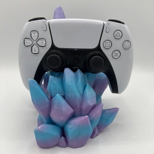 May include: A white PlayStation 5 controller rests on a 3D-printed crystal-shaped stand. The stand features a gradient of blue and purple hues. The controller has black accents and the PlayStation logo.