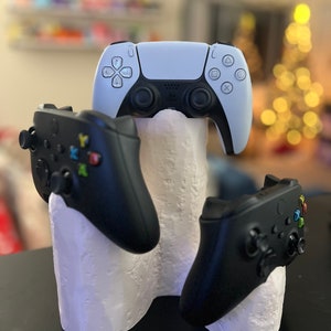 Może przedstawiać: Biały plastikowy stojak z trzema miejscami na kontrolery do gier wideo. Biały kontroler PlayStation 5 znajduje się w górnym miejscu, a dwa czarne kontrolery Xbox znajdują się w dolnych miejscach.
