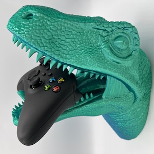 May include: A teal green dinosaur head wall mount with an open mouth holding a black video game controller.