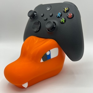 Puede incluir: Un soporte de impresión 3D en forma de personaje Pokémon, Charmander, en naranja y negro, sosteniendo un mando Xbox negro.