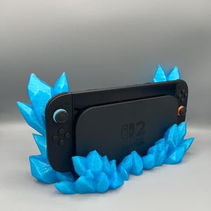 May include: A black Nintendo Switch console sits in a stand made of bright blue crystal shapes. The stand has multiple crystal formations. The console has a black screen and controllers with blue and orange accents. The words "NINTENDO SWITCH" are visible.