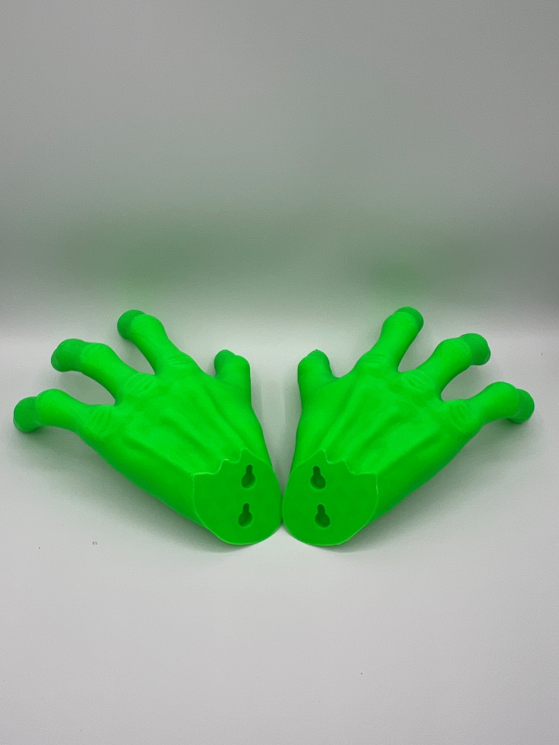 May include: A pair of bright green, three-fingered hand sculptures. Each hand features a textured, claw-like appearance with prominent knuckles and veins. The base of each hand is flat and includes two integrated keyhole slots for wall mounting.